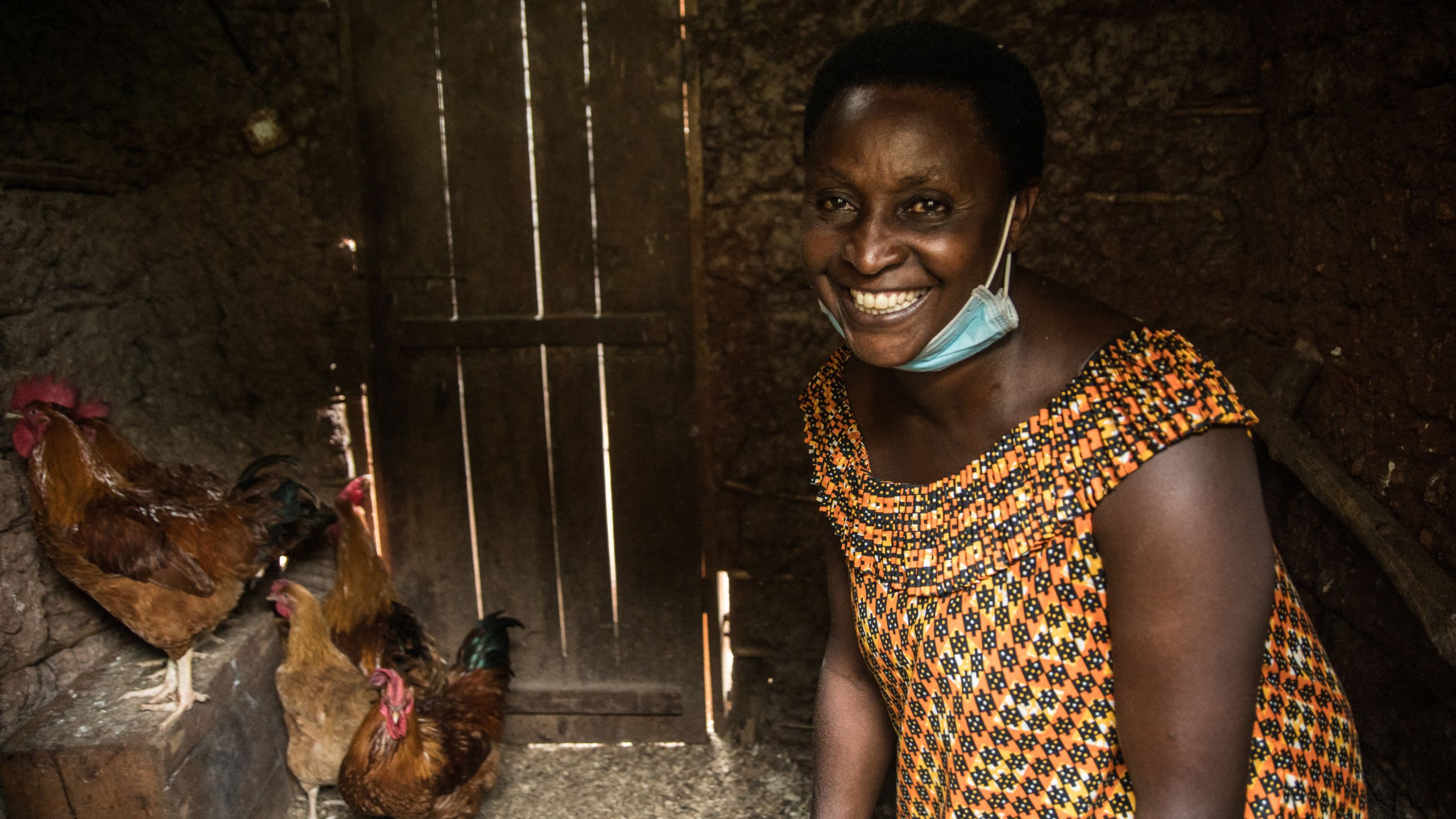 A smiling woman in an orange patterned dress stands inside a chicken coop with hens visible in the background.
