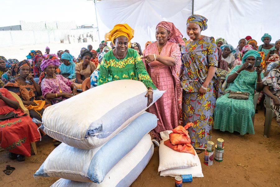 Three women stand next to three large white bags stuffed with seeds.